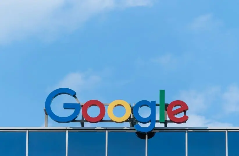 The Google logo in vibrant colors is displayed on a rooftop against a blue sky. The modern, iconic design conveys technology and innovation.