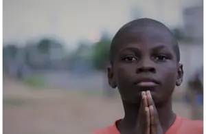 A young boy in an orange shirt stands outdoors with hands pressed together in a prayer-like gesture. The background is blurred, giving a serene atmosphere. Lent