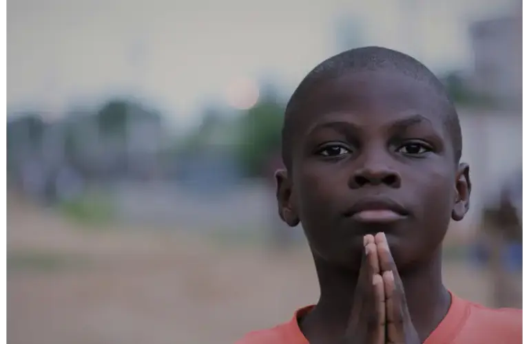 A young boy in an orange shirt stands outdoors with hands pressed together in a prayer-like gesture. The background is blurred, giving a serene atmosphere. Lent