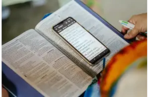 An open Bible, book, with a smartphone on top displaying text, held by a person with a pen in hand, indicating study or research, at church, home, or coffee shop. Bright and focused tone.