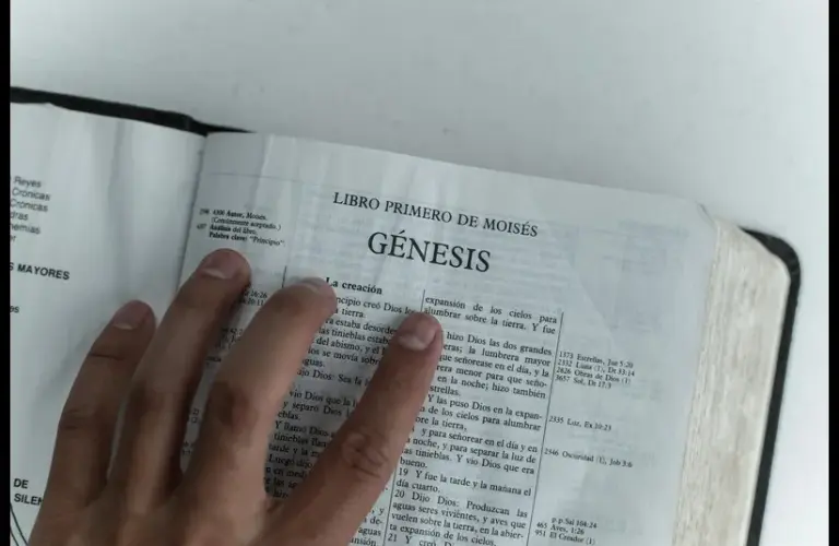 In daily devotion, a hand is placed on an open Bible on Genesis.