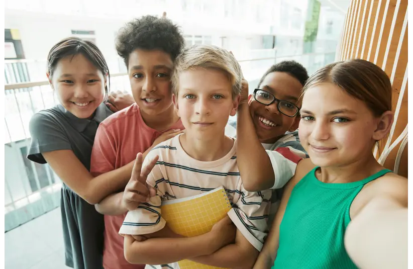 A diverse group of five smiling children stand closely, taking a selfie indoors. They exude joy and friendship, one holding a yellow notebook.