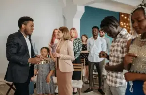 A group of people stand together in church, in a prayer or meditation setting. A man in a suit holds a collection plate. The mood is serene and focused. Group may include family and friends.