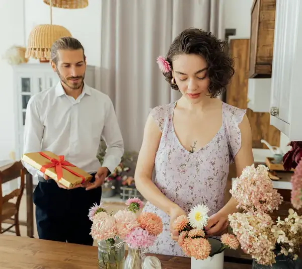 A woman in a floral dress arranges flowers on a kitchen table, while a man stands nearby holding a gift box for Valentine's Day. The setting is cozy and intimate.