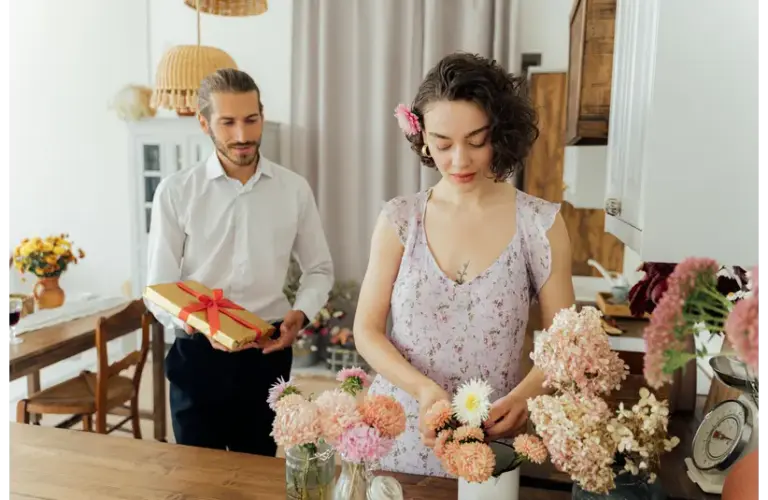 A woman in a floral dress arranges flowers on a kitchen table, while a man stands nearby holding a gift box for Valentine's Day. The setting is cozy and intimate.