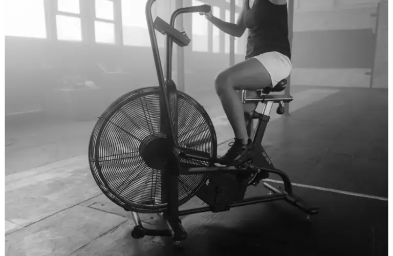 A person pedals an exercise bike with a large front fan in a spacious gym, creating a powerful, focused workout atmosphere as light filters through the windows.