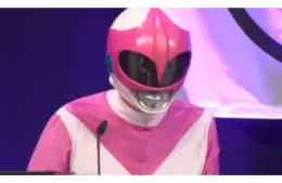 A person dressed in a pink and white superhero costume with a glossy helmet speaks at a podium. The background is a deep blue with partial lighting.