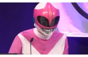 A person dressed in a pink and white superhero costume with a glossy helmet speaks at a podium. The background is a deep blue with partial lighting.