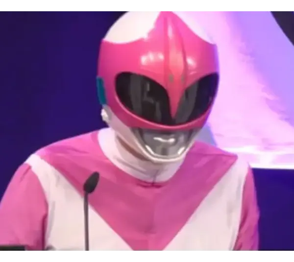 A person dressed in a pink and white superhero costume with a glossy helmet speaks at a podium. The background is a deep blue with partial lighting.