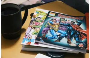 A stack of colorful comics featuring superheroes lies on a table next to a black coffee mug. The scene feels cozy and nostalgic.