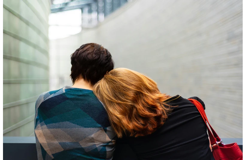 Two people sit closely, one resting their head on the other's shoulder, creating a sense of comfort and intimacy. They face away in a modern hallway.