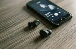A smartphone playing music with black wireless Google fast pair earbuds beside it on a wooden surface. The scene suggests a relaxed, tech-savvy vibe.