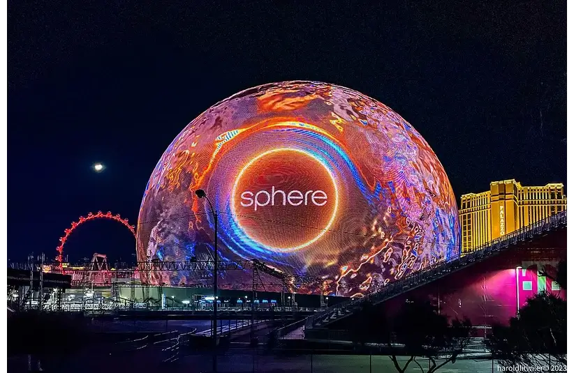 Vibrant dome in Vegas night sky, displaying "sphere" in glowing orange. Nearby, buildings illuminated, creating a lively, futuristic ambiance.