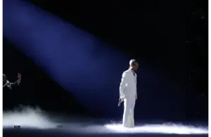 Pharrell Williams, in a white suit and sunglasses, stands in a spotlight on a smoky stage, holding a microphone. Another figure gestures in the dimly lit background.