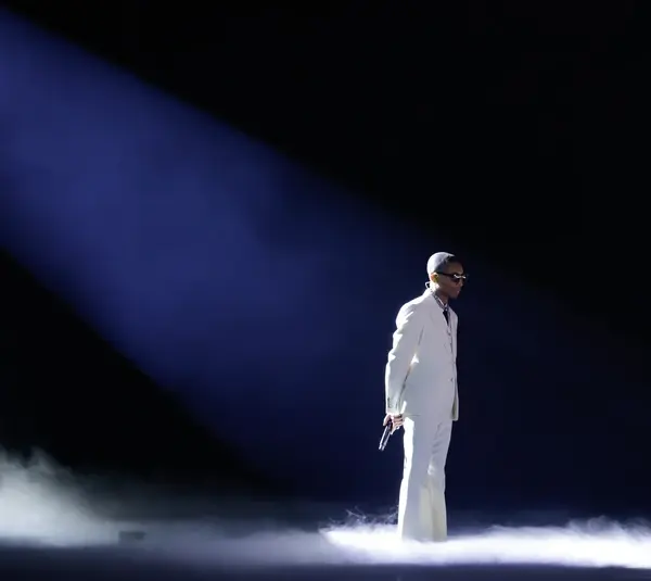 Pharrell Williams, in a white suit and sunglasses, stands in a spotlight on a smoky stage, holding a microphone. Another figure gestures in the dimly lit background.