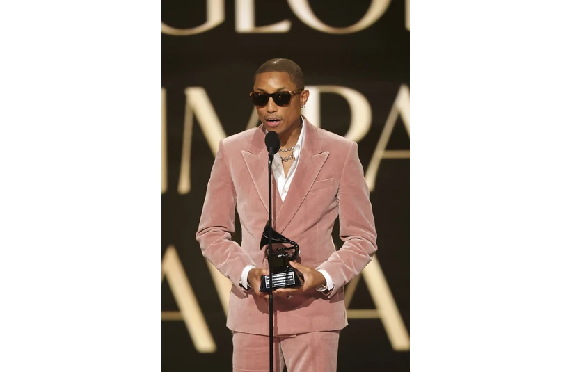 Pharrell Williams, in a pink velvet suit and sunglasses, speaks into a microphone on stage, holding an award. The background features large, out-of-focus text.