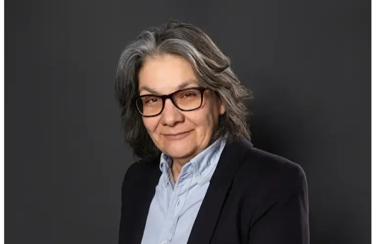 Ann Godoff, legendary editor, a person with gray hair and glasses smiles slightly. They wear a light blue shirt and dark blazer, set against a plain dark background, conveying warmth and professionalism.