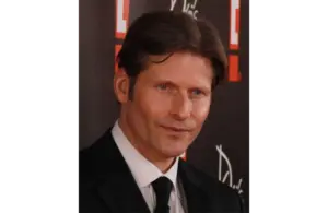 An image of Crispin Glover in a black suit and tie is smiling slightly against a dark backdrop with red and white patterns, creating a formal and composed mood.