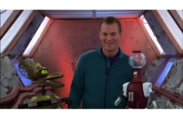 Mike Nelson, in a teal jumpsuit, smiles warmly between two robots, Tom Servo (on the right) and Crow T. Robot (on the left), on the Satellite of Love spaceship setting for MST3K. The mood is playful and light-hearted.