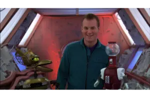 Mike Nelson, in a teal jumpsuit, smiles warmly between two robots, Tom Servo (on the right) and Crow T. Robot (on the left), on the Satellite of Love spaceship setting for MST3K. The mood is playful and light-hearted.