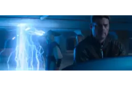 Three people, in Mortal Kombat 2, stand in a dimly lit room. Blue lightning arcs from the ceiling. The mood is tense, with focus on a man in a leather jacket in the foreground.