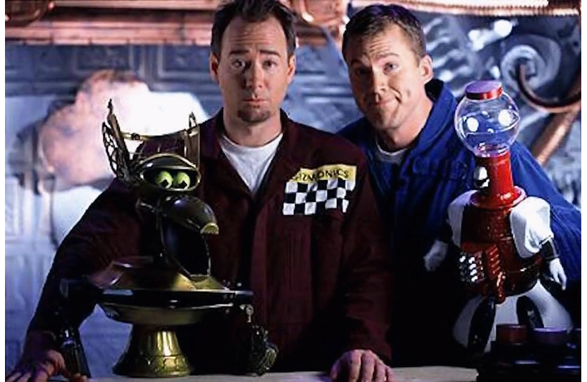 MST3K's Mike J. Nelson and Joel Hodgson stand with Crow T. Robot & Tom Servo on a spaceship set. One wears a maroon uniform with a checkered pattern, and the other is in blue. The mood is playful.