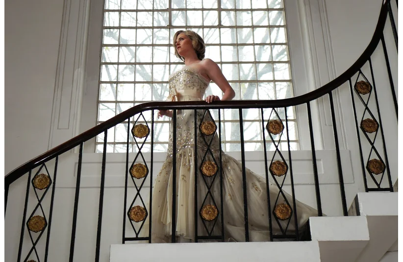 Kristen Stewart as Diana, Princess of Wales, in an elegant, bejeweled gown, stands on a curved staircase, bathed in light from a large window. The scene conveys elegance and poise.