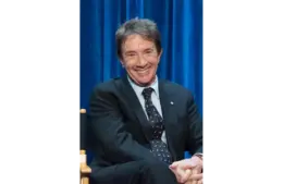 Martin Short, father of the late Katherine H. Short, sits in a suit sits smiling against a vibrant blue curtain backdrop. Their relaxed posture and cheerful expression convey a friendly, welcoming tone. The Short family is in mourning.