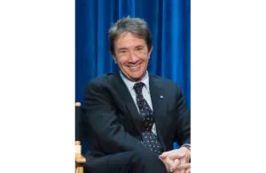 Martin Short, father of the late Katherine H. Short, sits in a suit sits smiling against a vibrant blue curtain backdrop. Their relaxed posture and cheerful expression convey a friendly, welcoming tone. The Short family is in mourning.