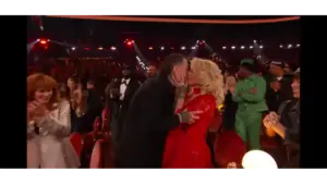 Jelly Roll and wife, Bunnie Xo, share a kiss in a crowded theater, surrounded by applauding people. The woman is in a red dress and the man in a black jacket. The atmosphere is joyful and celebratory.