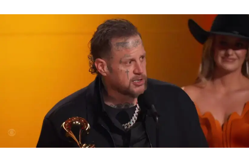 Jelly Roll holds a trophy and speaks into a microphone on stage. A woman in an orange outfit and black hat smiles in the background.