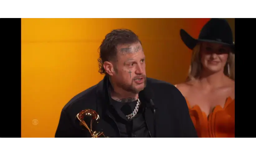 A man, Jelly Roll, with tattoos on his face, speaks into a microphone while holding a trophy against a bright orange background. A woman in a cowboy hat stands smiling behind him. The mood appears celebratory.