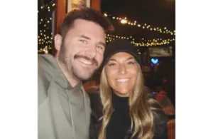 Screenshot of Brianna “Bri” McNees and Connor Spies, Courtesy of breezy_mcneezy via Instagram, shows a smiling man and woman pose for a selfie in a dimly lit room with string lights in the background, creating a warm and cheerful atmosphere.