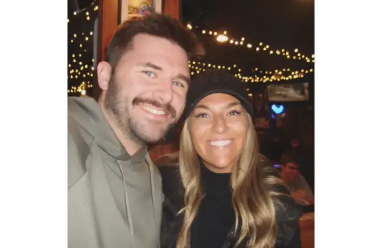 Screenshot of Brianna “Bri” McNees and Connor Spies, Courtesy of breezy_mcneezy via Instagram, shows a smiling man and woman pose for a selfie in a dimly lit room with string lights in the background, creating a warm and cheerful atmosphere.