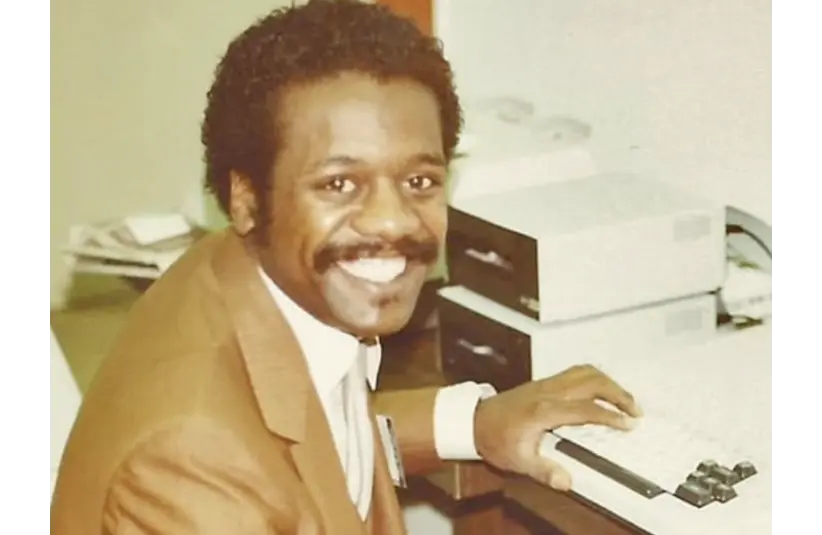 Edward Smith, creator of the first pc-like gaming system, the imagination machine is  in a brown suit sits at a vintage computer, typing. The scene conveys a positive, professional atmosphere in an office setting.