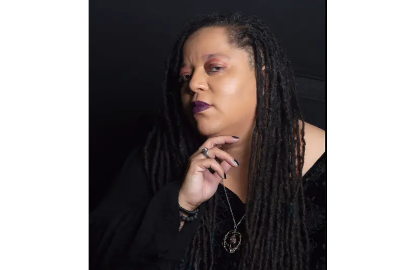 Portrait of a Tanya DePass, a black gamer, streamer and minority advocate, with long dreadlocks, wearing dark clothing and a pendant. They look thoughtfully at the camera with a confident expression.