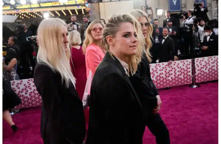 Kristen Stewart, who is restoring the Historic Highland Theatre, walks with a group of people in stylish attire pose on a vibrant pink carpet. They are surrounded by photographers against a backdrop of bright lights and cameras, exuding a lively, celebratory mood.