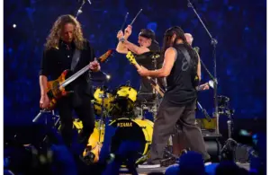 Metallica, a rock band performs energetically on stage. One guitarist is strumming intensely, another faces the drummer, who is mid-action. Their backdrop is a vast, cheering crowd.