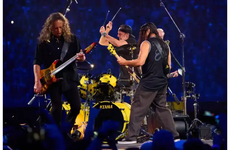 Metallica, a rock band performs energetically on stage. One guitarist is strumming intensely, another faces the drummer, who is mid-action. Their backdrop is a vast, cheering crowd.