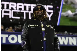 Lil Jon stands smiling at a stadium, wearing sunglasses, a Baltimore Ravens jacket, and a beanie. The background shows a blurred stadium sign. The mood is energetic.