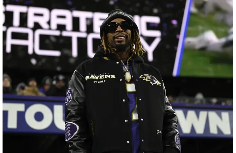 Lil Jon stands smiling at a stadium, wearing sunglasses, a Baltimore Ravens jacket, and a beanie. The background shows a blurred stadium sign. The mood is energetic.