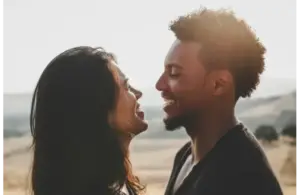 A couple stands closely, smiling at each other against a sunlit, blurred countryside. The mood is warm and joyful, capturing an intimate, happy moment, emotional clarity