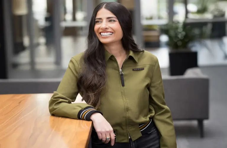 Smiling woman, Asha Sharma, with long dark hair leans against a wooden table, wearing an olive green jacket. Office setting with a relaxed, cheerful atmosphere.