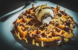 image of A warm waffle drizzled with chocolate sauce, topped with a scoop of vanilla ice cream and sprinkled with crushed nuts, sits on a white plate. Indulgent and inviting for Galentine's Day brunch