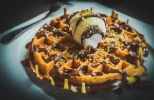 image of A warm waffle drizzled with chocolate sauce, topped with a scoop of vanilla ice cream and sprinkled with crushed nuts, sits on a white plate. Indulgent and inviting for Galentine's Day brunch