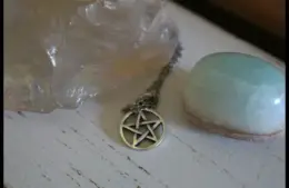 A silver pentacle necklace lies between a large translucent crystal and a smooth, light blue stone on a rustic white wooden surface, conveying a mystical and serene atmosphere. Wiccan Rede