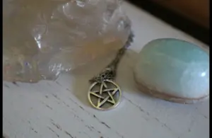 A silver pentacle necklace lies between a large translucent crystal and a smooth, light blue stone on a rustic white wooden surface, conveying a mystical and serene atmosphere. Wiccan Rede
