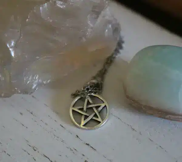 A silver pentacle necklace lies between a large translucent crystal and a smooth, light blue stone on a rustic white wooden surface, conveying a mystical and serene atmosphere. Wiccan Rede