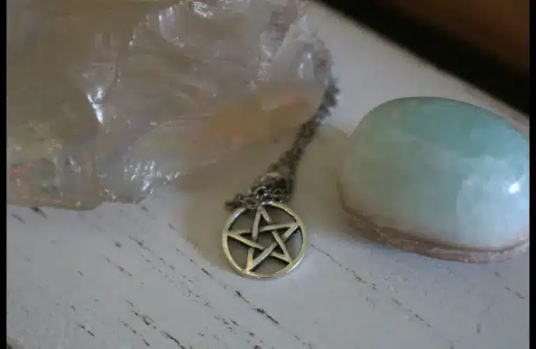 A silver pentacle necklace lies between a large translucent crystal and a smooth, light blue stone on a rustic white wooden surface, conveying a mystical and serene atmosphere. Wiccan Rede