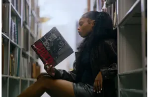 Young woman in a leather jacket sits on a library floor, reading a magazine titled "Rodeo" for Black History Month. She appears focused, surrounded by tall shelves of books.